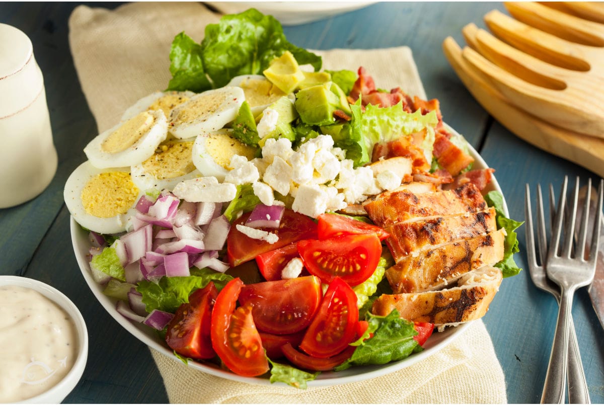 A white bowl filled with Cobb Salad (grilled chicken, tomato, red onion, cheese, boiled egg, avocado, bacon & greens).