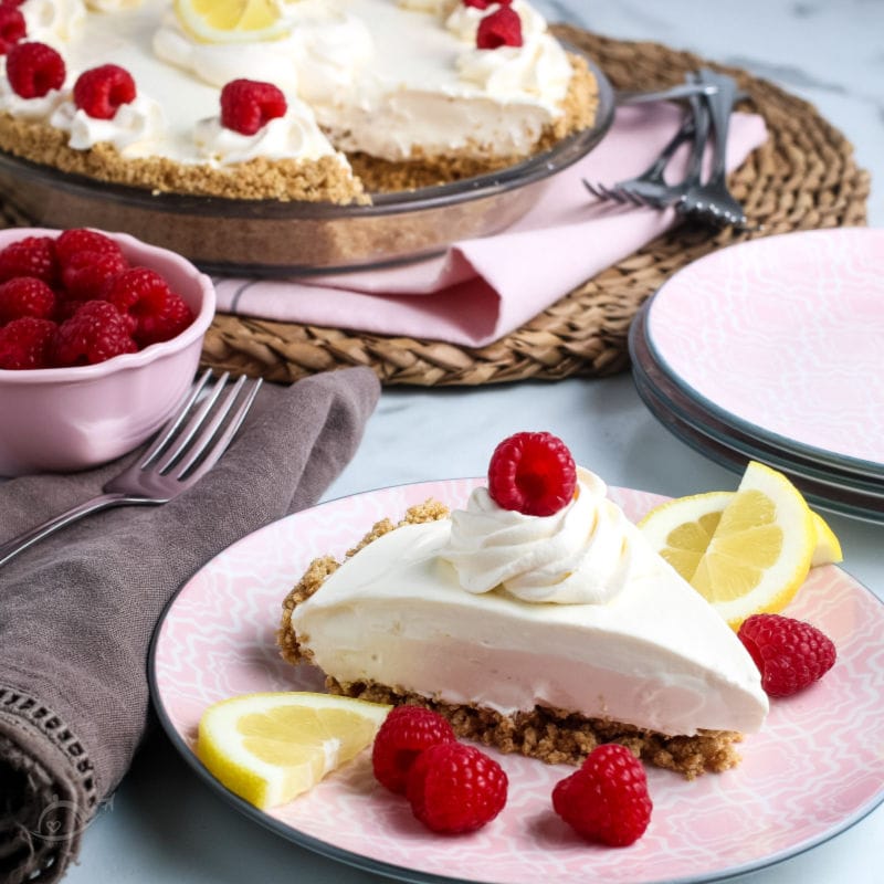 A slice of Lemonade Pie garnished with whipped cream, berries & lemon slices on a pink plate. A bowl of raspberries & partial pie in the background | A Reinvented Mom