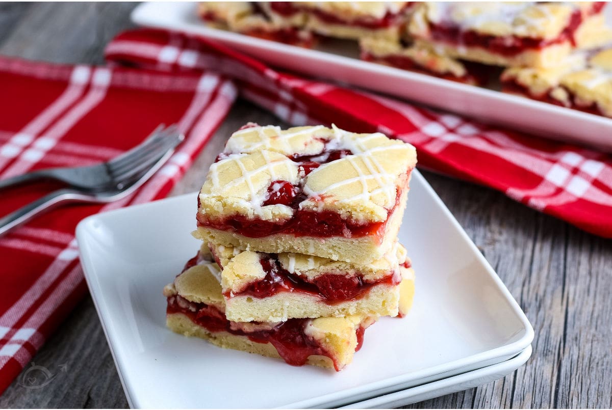 White plate with three Cherry Pie Bars on a wood background with red & white napkins, 2 forks & serving plate of cherry bars. | A Reinvented Mom