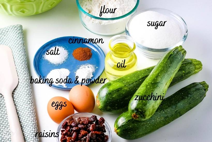 all ingredients needed for zucchini bread muffins on white counter next to hand towel and spatula