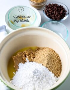 Powdered sugar, peanut butter, graham crackers, and butter in a white bowl for the first layer of peanut butter squares.