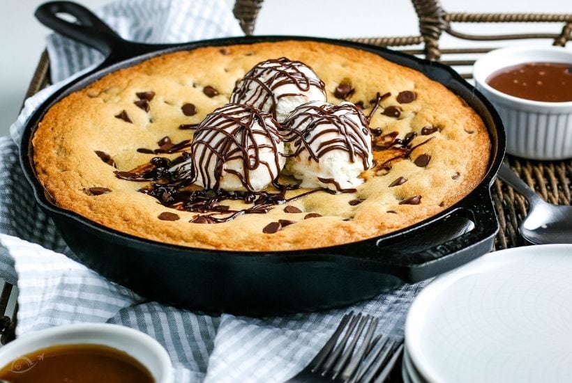A skillet cookie in a cast iron pan after baking topped with vanilla ice cream and chocolate syrup.