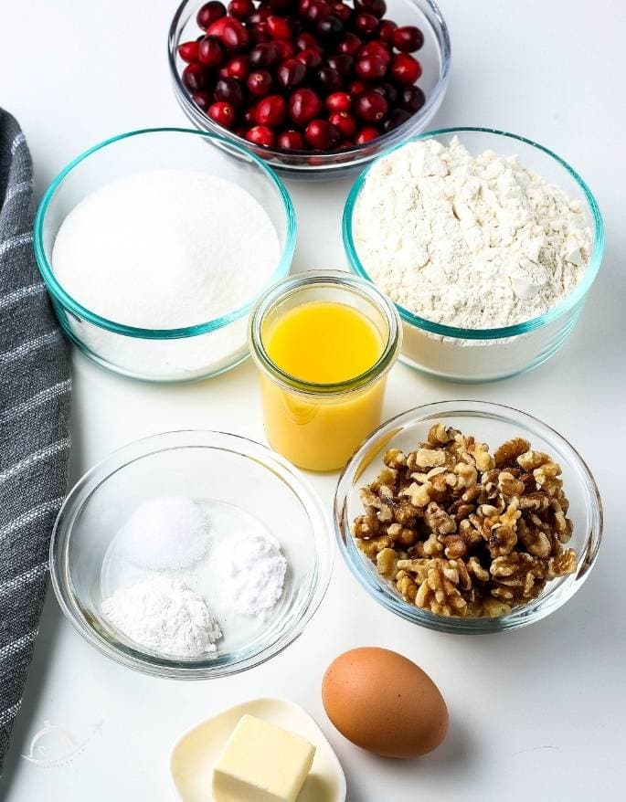 ingredients for orange cranberry bread in glass bowls on white counter.