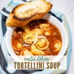 White bowl filled with tortellini soup & a slice of bread with striped towel and graphic overlay.