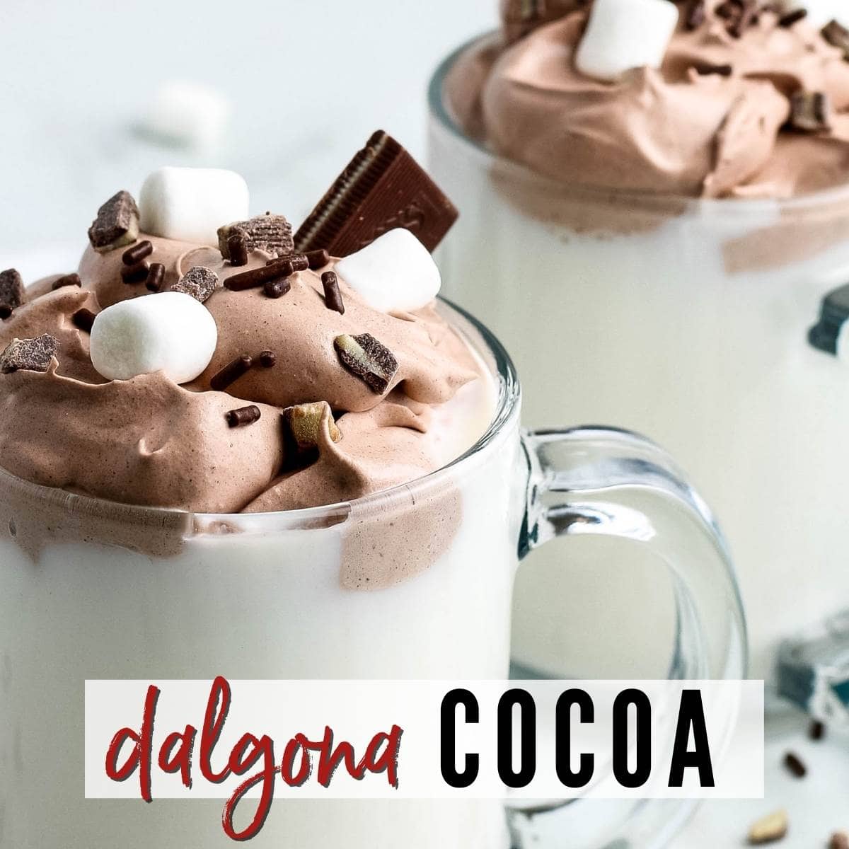 Two mugs of Dalgona whipped cocoa garnished with jimmies & marshmallows with a graphic overlay