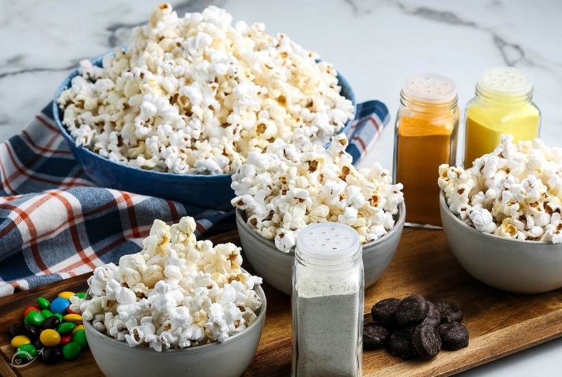 Four bowls of popcorn, different containers of seasoning, M&M candy and chocolate candy placed on wooden board