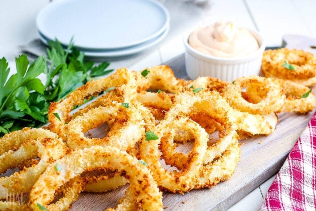 homemade air fried onion rings on a cutting board with sauce and garnish