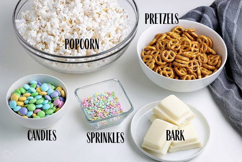 Bowls filled with ingredients for Easter snack mix on white counter with text overlay.