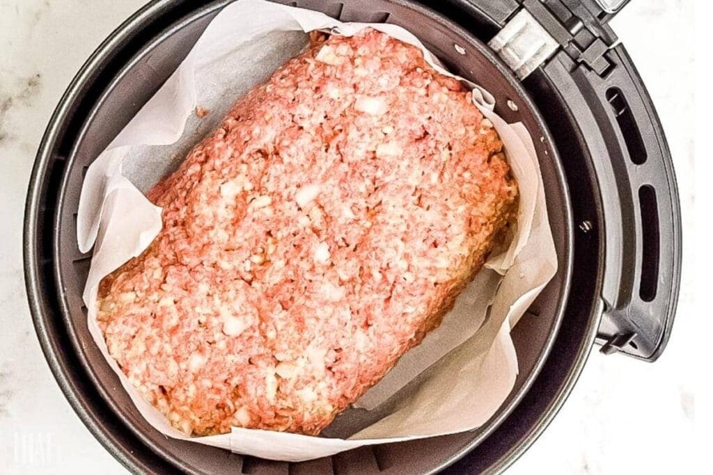 Raw shaped meatloaf on parchment paper in air fryer basket.