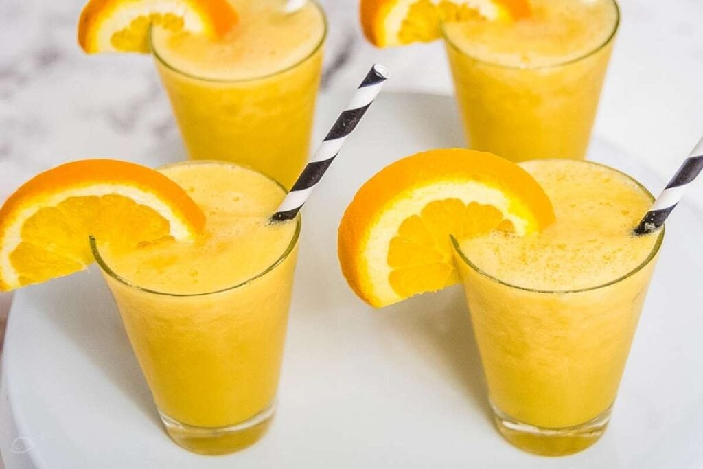 closeup view of four glasses of blended drinks with straws and orange slice garnishes on a white background