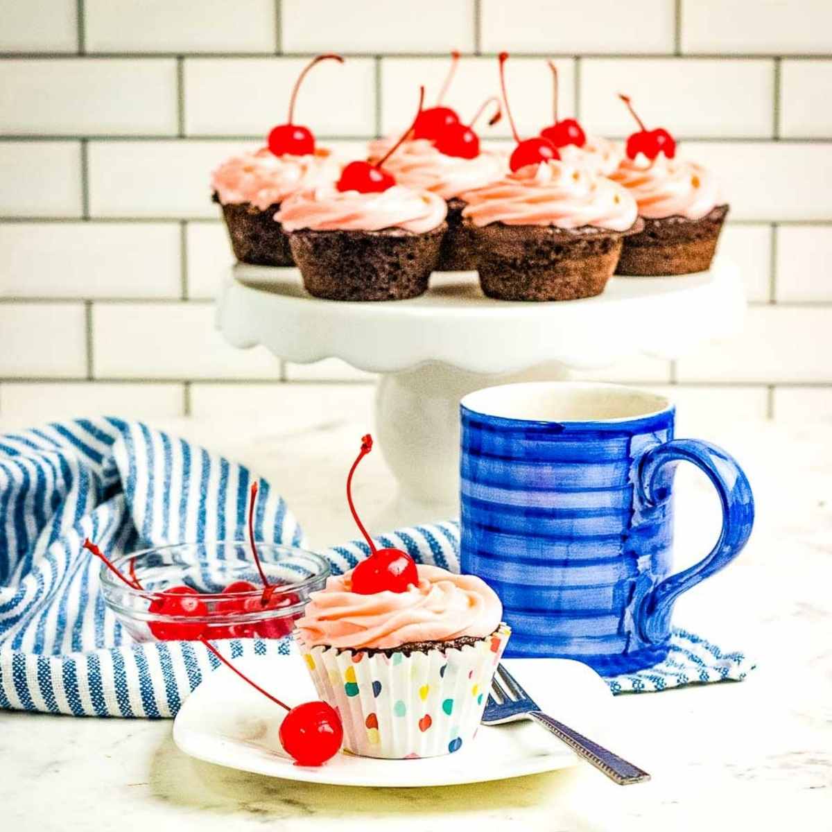 baked cherry chocolate dr. pepper cupcakes on white plates with a blue coffee mug and napkin in background