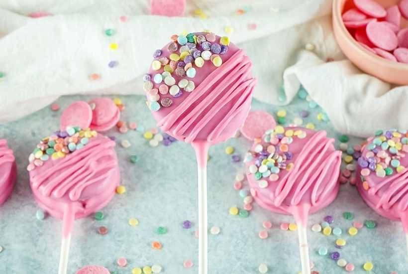 Close up of valentines day oreo pops with sprinkles and pink chocolate wafers in the background.