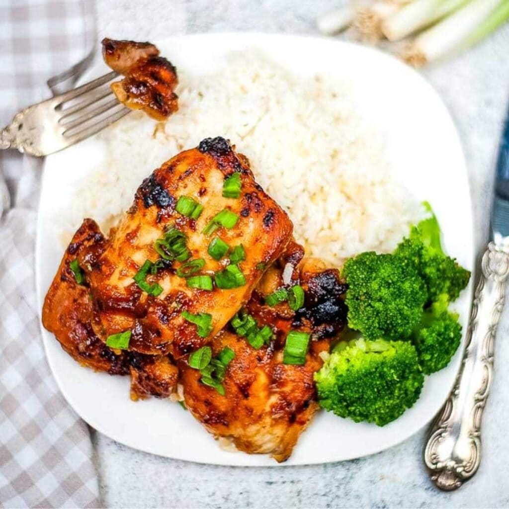 Two pieces of Air Fryer Huli Huli Chicken on a plate with broccoli and white rice next to fork and knife.