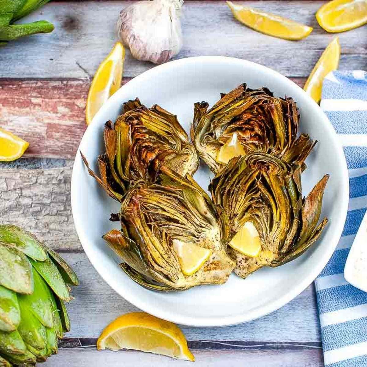 air fryer artichokes in a small white bowl next to lemon wedges and a whole artichoke