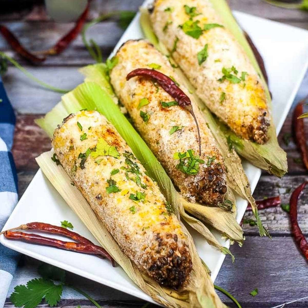 three ears of air fried Mexican street corn on the husk on a white plate