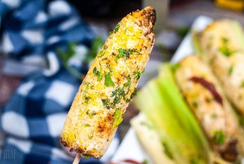 holding a skewer of mexican street corn covered in cheese and parsley