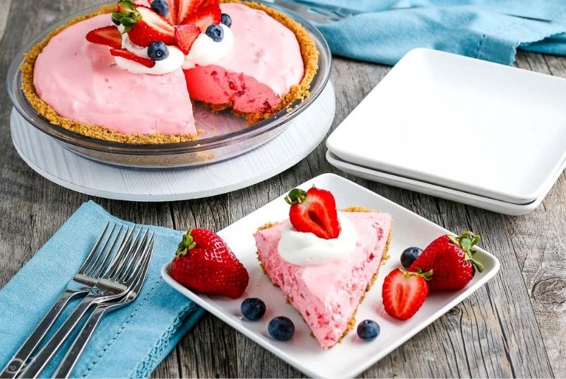 A slice of creamy jello pie garnished with berries and Cool Whip whipped topping on a white plate with full pie, napkin, cutlery & plates in the background.