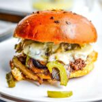 Closeup view of a Philly cheesesteak slider with ground beef on a white plate with peppers, mushrooms and cheese spilling out.