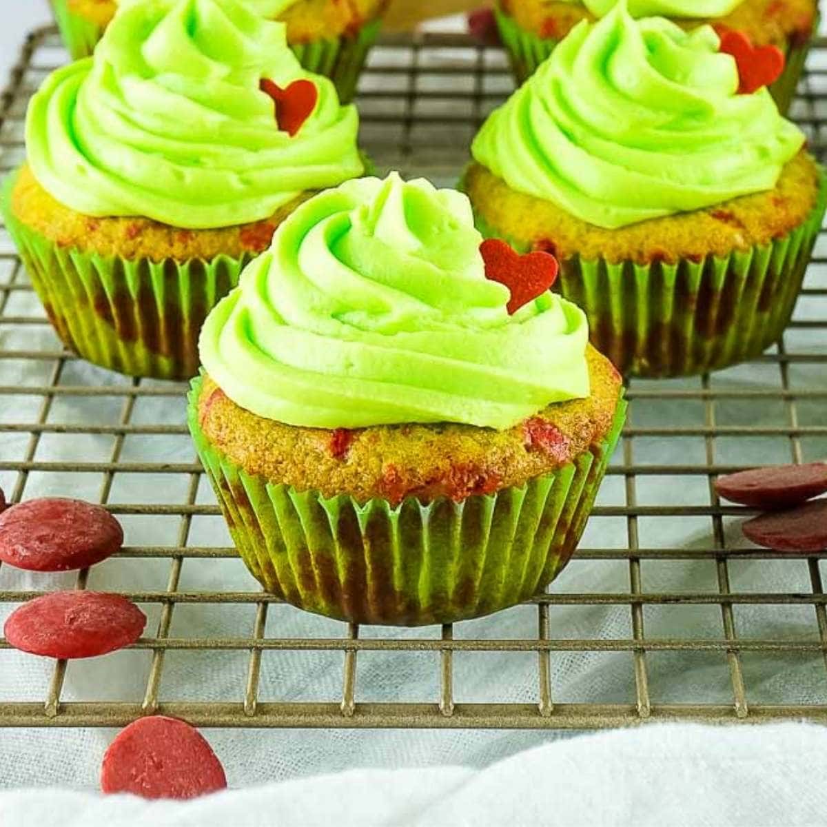 a close up of grinch cupcakes with small red hearts sitting on a cooling rack.