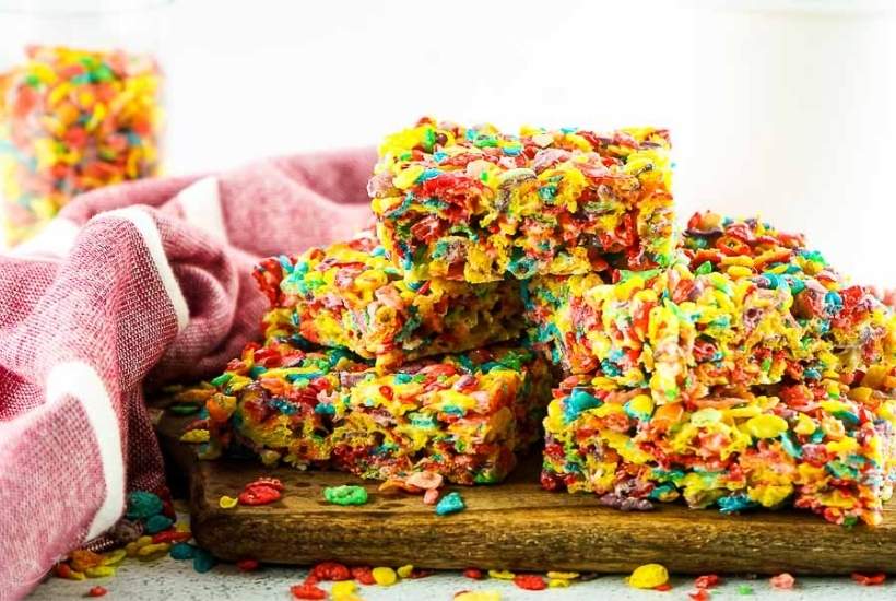 a stack of fruity pebble rice krispie treats cut into squares on a wood cutting board.