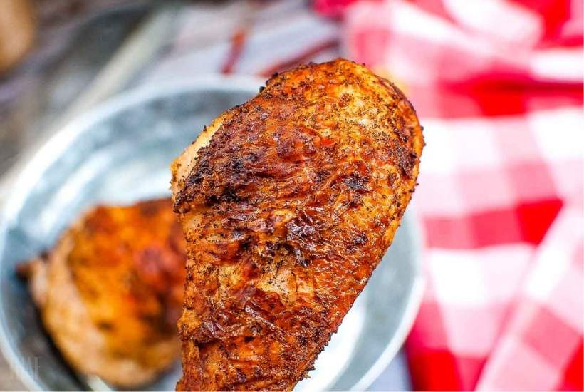 a close up of an air fried turkey leg with another turkey leg in a metal pan in the background next to a red and white plaid table cloth.