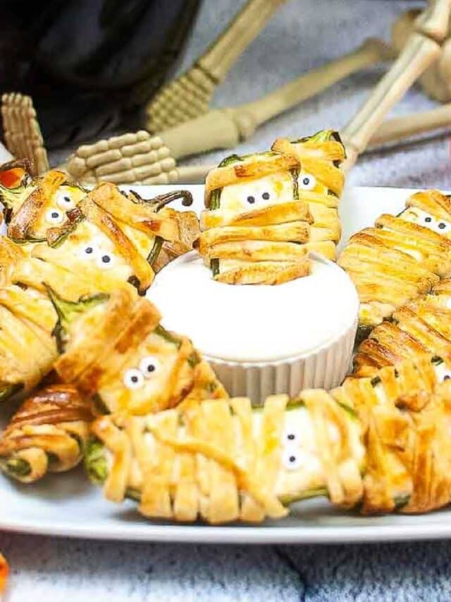 entire plate of mummy jalapeno poppers on a square plate one dipped in white sauce in the center of the plate