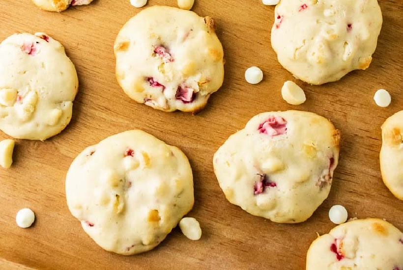 Top view of strawberry cream cheese cookies on a wood cutting board with white chips sprinkled around them.