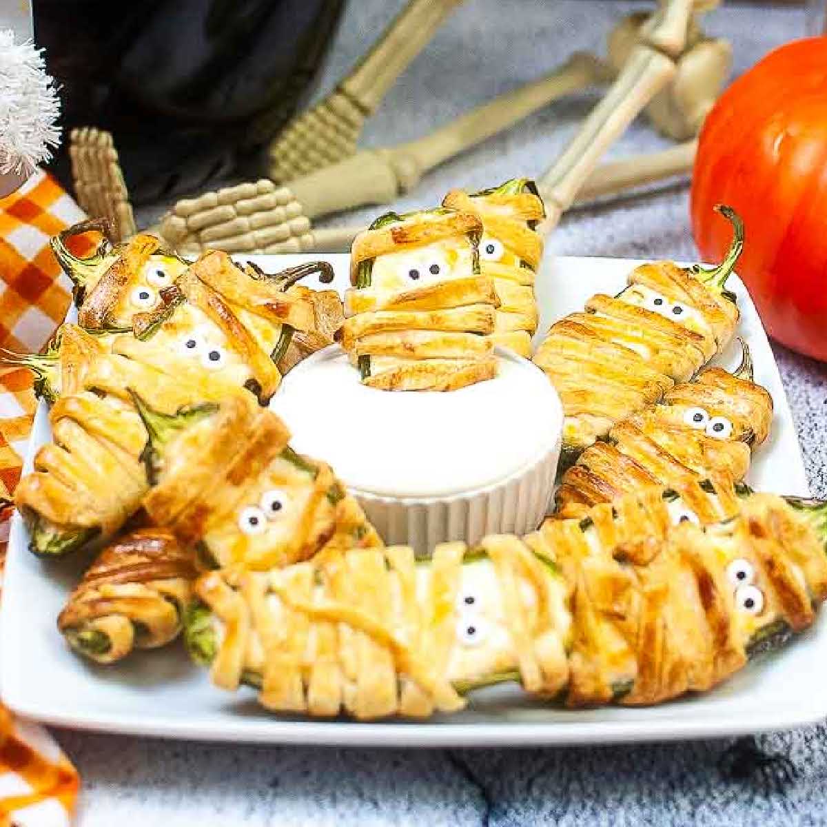 square white plate filled with jalapeno popper mummies and one dipped in white sauce in the center of the plate.