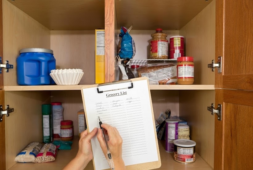 Hands holding a grocery list on a clipboard in front of an open kitchen cabinet.