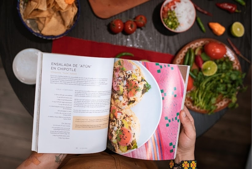 Hands holding an open cookbook with food in the background.