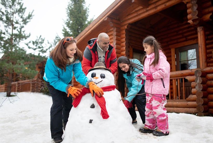 family building a snowman outside of a wood cabin.