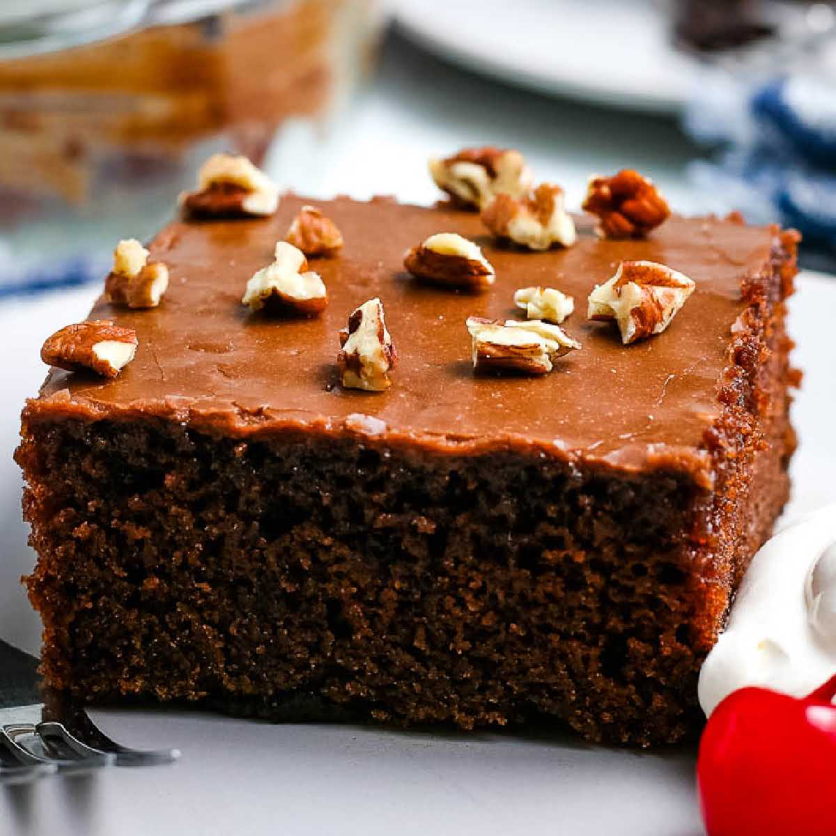 A slice of old fashioned Coca Cola cake topped with chopped nuts next to a cherry and whipped cream.
