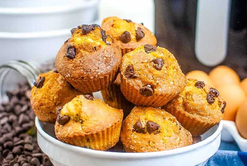 chocolate chip muffins stacked in a white bowl with chocolate chips and eggs in the background.
