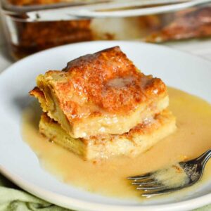 two slices of old fashioned bread pudding with vanilla sauce on a white plate with a fork.