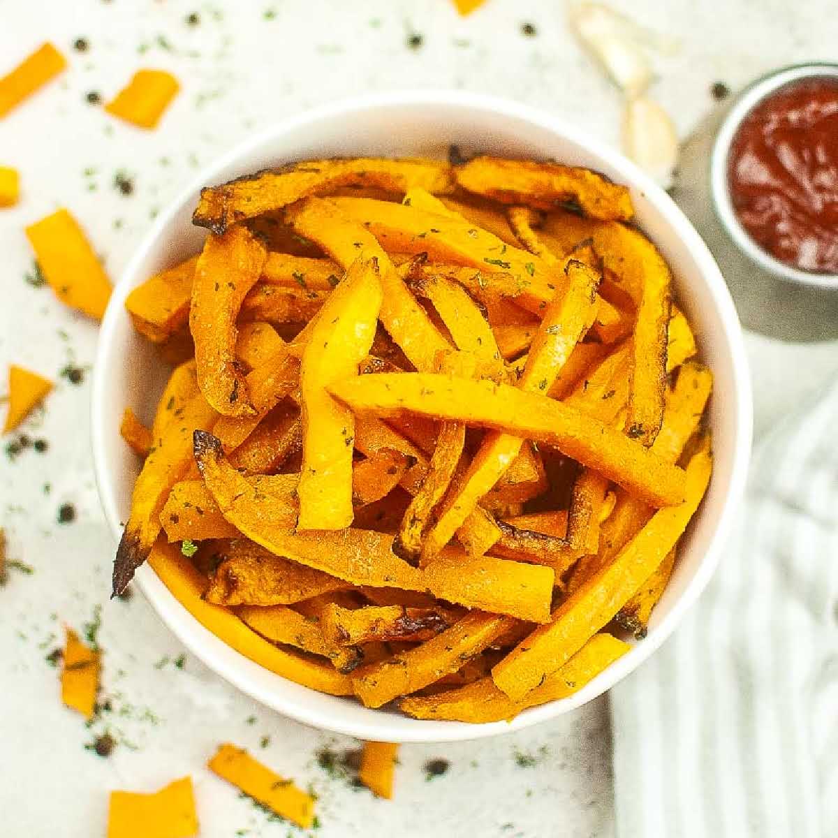 top view of butternut squash fries in a white bowl next to a bowl of ketchup.