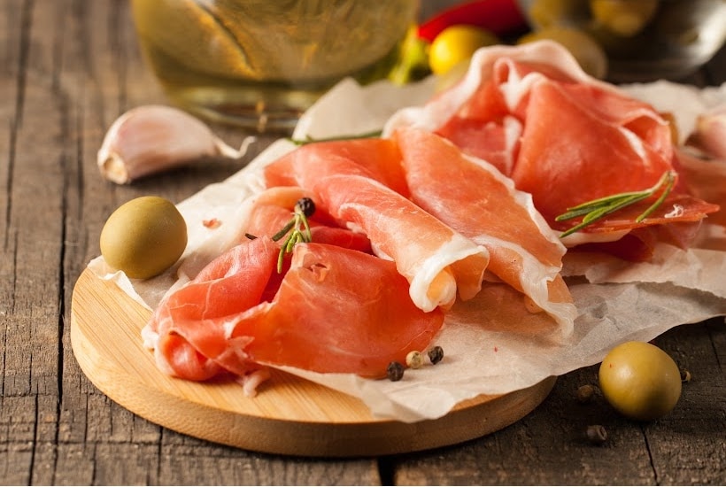 very thinly sliced ham on paper with olives on a round wooden cutting board.