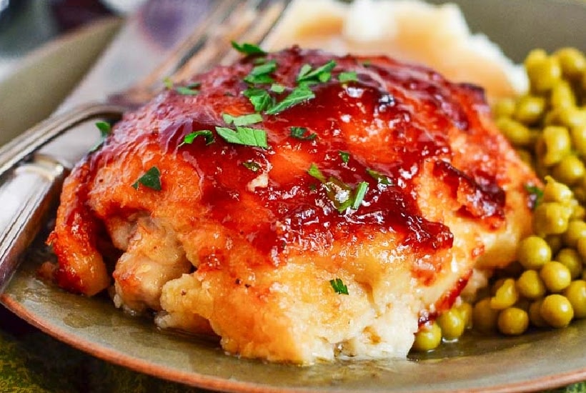 Closeup view of oven baked BBQ chicken thighs on a plate with peas, mashed potatoes, and a fork.