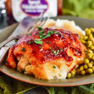 Front view of a plate with BBQ Baked Chicken Thighs, peas, mashed potatoes and a fork and knife.