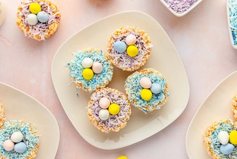 square plate of four Rice Krispie nests with mini eggs surrounded by additional nests and ingredients.