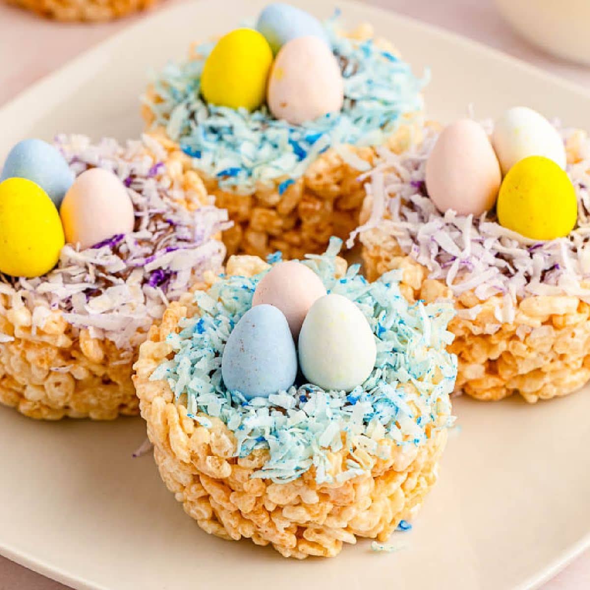 closeup view of Rice Krispie nests with dyed coconut and mini chocolate eggs on a white plate.
