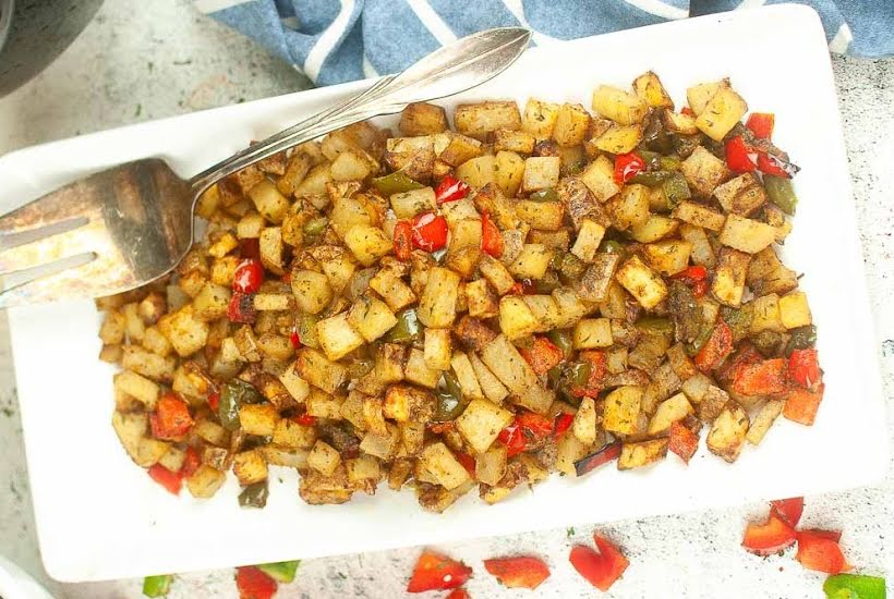 Crispy breakfast potatoes and peppers on a white rectangular plate with a large serving fork.