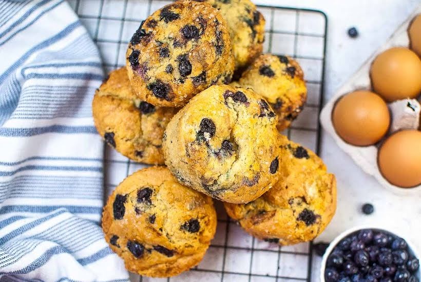 Overhead view of air fried blueberry muffins on a wire cooling rack next to eggs and a bowl of blueberries.