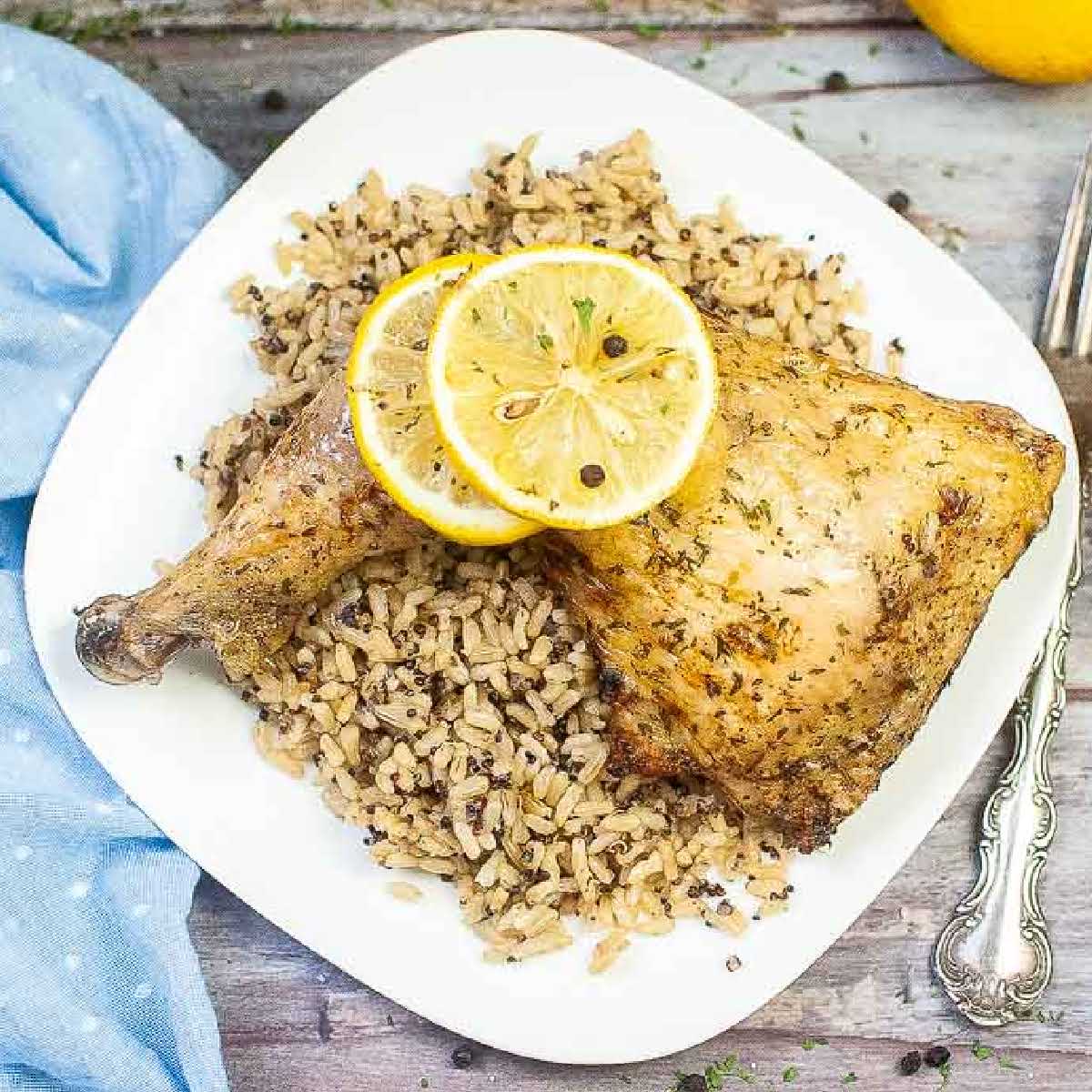 Overhead view of an air fried chicken leg quarter on a white plate with wild rice and lemon slices.