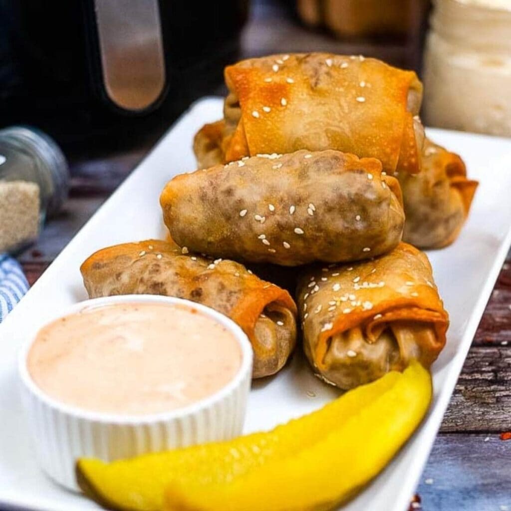 Air fried cheeseburger egg rolls stacked on a white tray with dipping sauce and pickle spears.