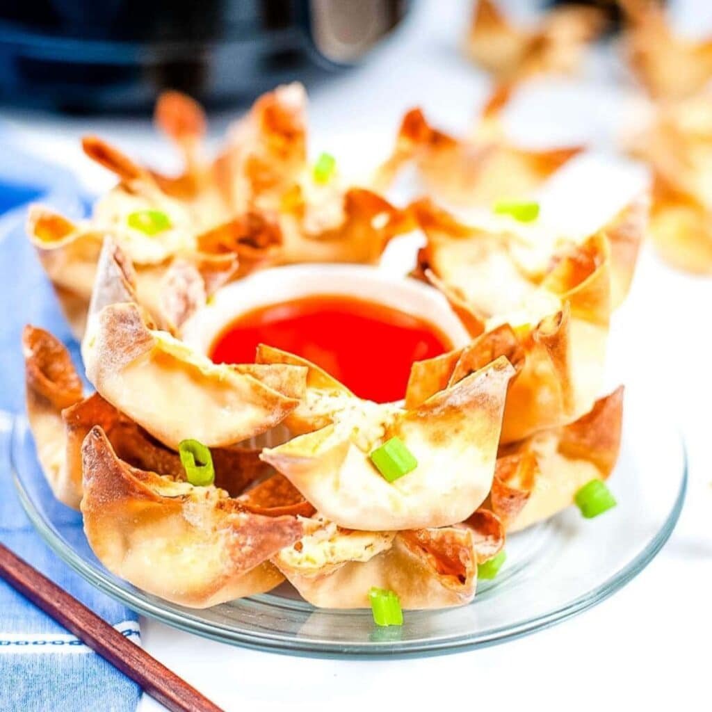 Air fried crab rangoon stacked on a glass plate with a bowl of sweet and sour dipping sauce.