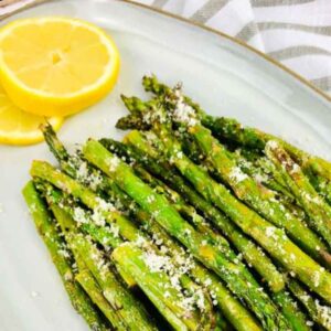 Air fried asparagus spears garnished with grated parmesan and lemon slices on a white platter.