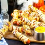 Mummy dogs on a wooden cutting board with a small bowl of mustard.