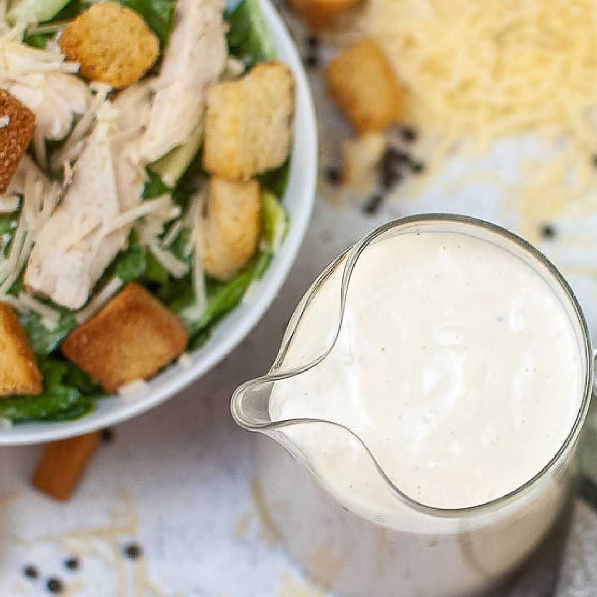 Top view of decanter of Caesar dressing without anchovies with chicken salad in the background.