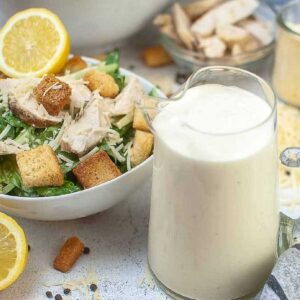 Clear decanter of Caesar dressing with a salad in the background.