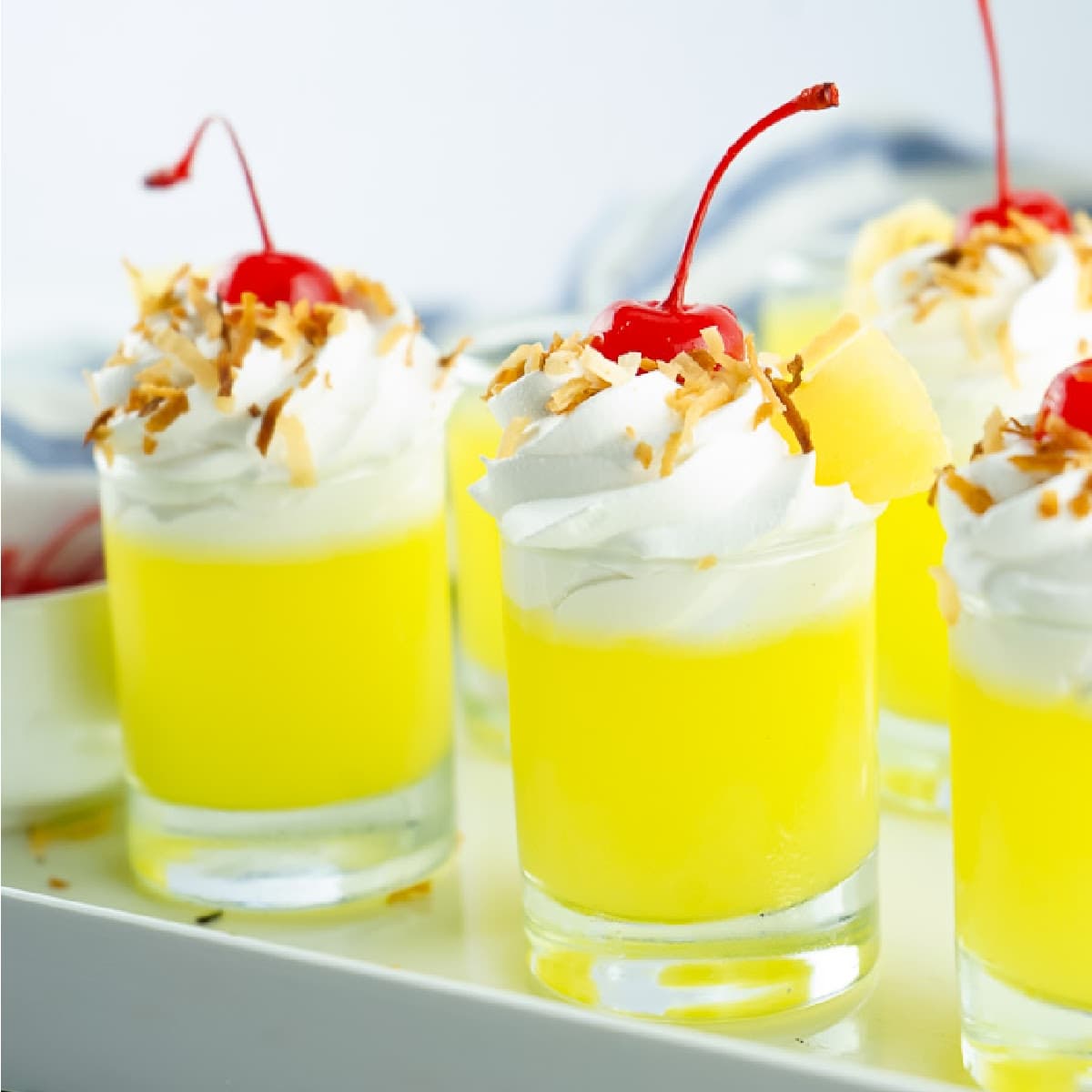 Pina Colada Jello Shots topped with whipped cream, toasted coconut, and cherries on a white tray.